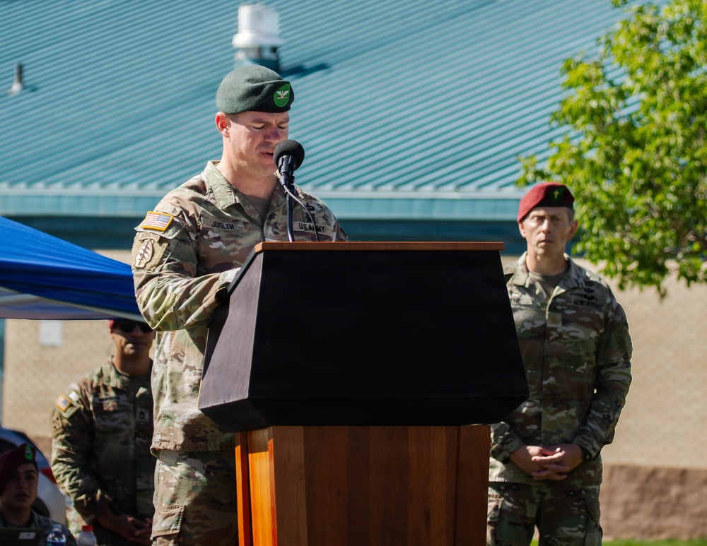 A U.S. Army soldier wearing a green beret is speaking at a podium during what appears to be an outdoor military ceremony. His name tape reads 'JOSLYN' and he wears the U.S. Army label with camouflage OCP uniform. Additional military personnel are visible in the background, including one wearing a maroon beret.
