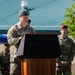 A U.S. Army soldier wearing a green beret is speaking at a podium during what appears to be an outdoor military ceremony. His name tape reads 'JOSLYN' and he wears the U.S. Army label with camouflage OCP uniform. Additional military personnel are visible in the background, including one wearing a maroon beret.
