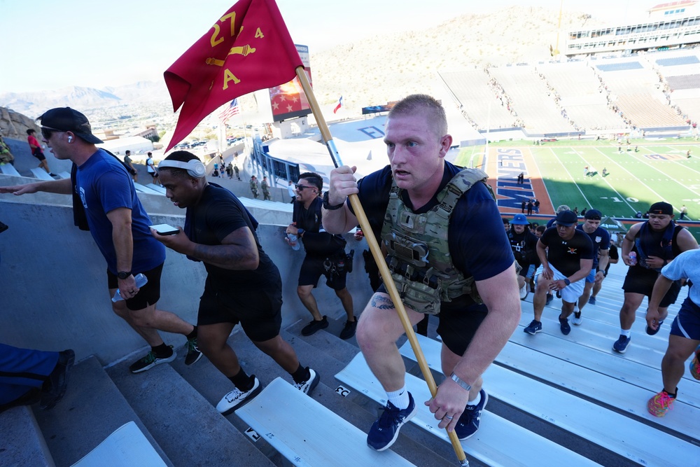 A group of participants are climbing stadium stairs during what appears to be a military-themed fitness challenge or memorial event. The lead participant is wearing a tactical plate carrier vest and carrying a red Marine Corps guidon flag with the number '27' visible. The event is taking place at a football stadium, identifiable by the green field and scoreboard visible in the background.