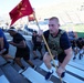A group of participants are climbing stadium stairs during what appears to be a military-themed fitness challenge or memorial event. The lead participant is wearing a tactical plate carrier vest and carrying a red Marine Corps guidon flag with the number '27' visible. The event is taking place at a football stadium, identifiable by the green field and scoreboard visible in the background.