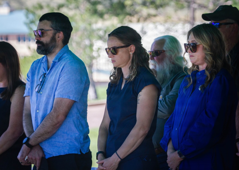A group of adults standing together outdoors in what appears to be a solemn or ceremonial gathering, possibly a memorial or formal event. Most individuals are wearing sunglasses and dark or blue-toned clothing, with serious, downward-cast expressions. The setting appears to be a park or institutional grounds with trees and a building visible in the background.