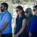 A group of adults standing together outdoors in what appears to be a solemn or ceremonial gathering, possibly a memorial or formal event. Most individuals are wearing sunglasses and dark or blue-toned clothing, with serious, downward-cast expressions. The setting appears to be a park or institutional grounds with trees and a building visible in the background.
