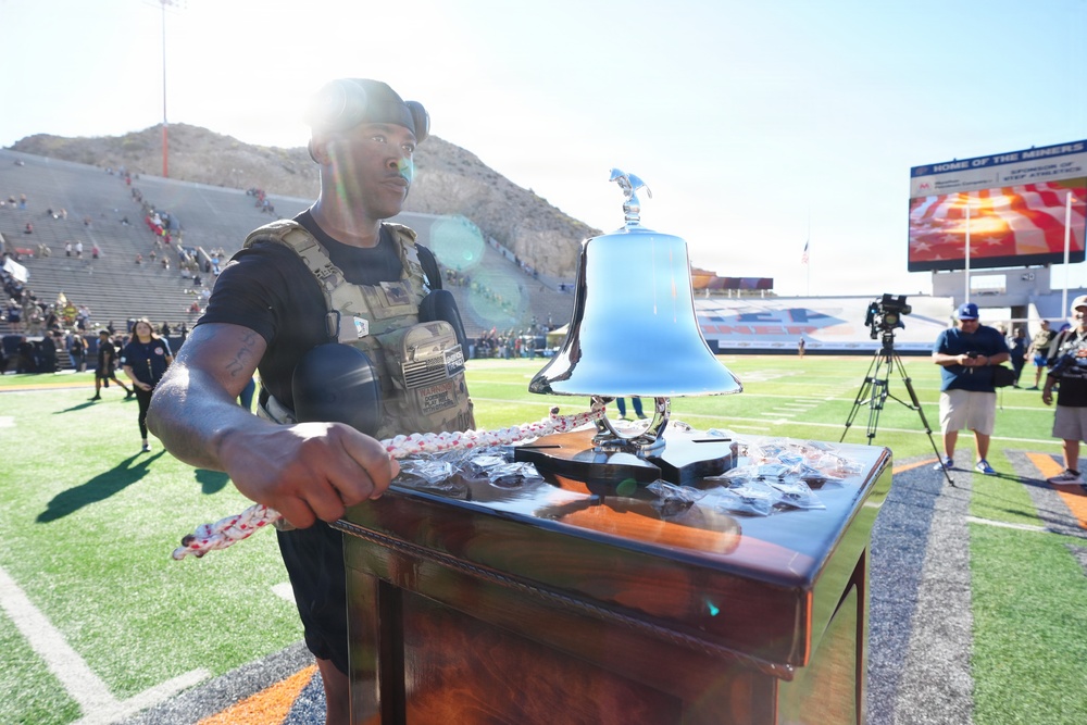 A muscular man wearing a tactical vest and black t-shirt stands at a wooden podium on a football field, holding a chain attached to a large decorative silver bell. The setting appears to be a pre-game or ceremonial event at a college football stadium, identifiable by the scoreboard text 'Home of the Miners,' suggesting UTEP (University of Texas at El Paso). A rocky mountain is visible in the background, consistent with the El Paso, Texas landscape.