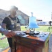 A muscular man wearing a tactical vest and black t-shirt stands at a wooden podium on a football field, holding a chain attached to a large decorative silver bell. The setting appears to be a pre-game or ceremonial event at a college football stadium, identifiable by the scoreboard text 'Home of the Miners,' suggesting UTEP (University of Texas at El Paso). A rocky mountain is visible in the background, consistent with the El Paso, Texas landscape.