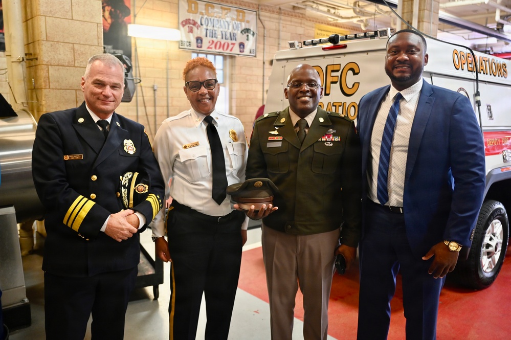 Four individuals pose for a group photo inside what appears to be a Washington D.C. fire station. The group includes a senior fire department official in dress uniform, a fire department officer in white shirt and black tie, a U.S. Army soldier in dress greens holding a military cap, and a civilian in a suit. A banner reading 'D.C.F.D. Company of the Year 2007' is visible in the background.