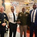 Four individuals pose for a group photo inside what appears to be a Washington D.C. fire station. The group includes a senior fire department official in dress uniform, a fire department officer in white shirt and black tie, a U.S. Army soldier in dress greens holding a military cap, and a civilian in a suit. A banner reading 'D.C.F.D. Company of the Year 2007' is visible in the background.