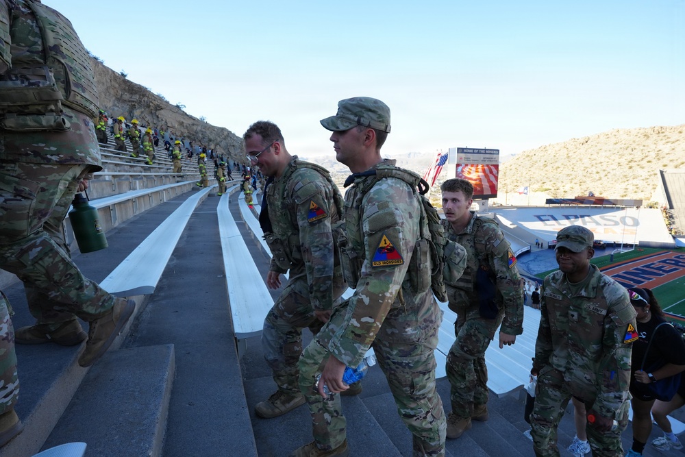 U.S. Army soldiers in OCP (Operational Camouflage Pattern) uniforms are climbing stadium steps at what appears to be the Sun Bowl stadium in El Paso, Texas. The soldiers are wearing tactical gear and backpacks, suggesting a physical fitness or memorial stair-climb event. In the background, firefighters in full gear are also visible climbing the stadium bleachers, indicating a joint commemorative event.
