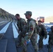 U.S. Army soldiers in OCP (Operational Camouflage Pattern) uniforms are climbing stadium steps at what appears to be the Sun Bowl stadium in El Paso, Texas. The soldiers are wearing tactical gear and backpacks, suggesting a physical fitness or memorial stair-climb event. In the background, firefighters in full gear are also visible climbing the stadium bleachers, indicating a joint commemorative event.