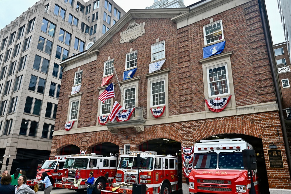 The image shows a historic brick fire station belonging to the DC Fire Department (D.C.F.D.), decorated with American flags, patriotic bunting, and military branch flags for what appears to be a ceremonial or commemorative event. Multiple red fire trucks are parked in front of the station bays, with unit numbers 13 and 16 visible. The building features colonial-style architecture with arched bays, multi-pane windows, and a central balcony.