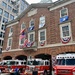 The image shows a historic brick fire station belonging to the DC Fire Department (D.C.F.D.), decorated with American flags, patriotic bunting, and military branch flags for what appears to be a ceremonial or commemorative event. Multiple red fire trucks are parked in front of the station bays, with unit numbers 13 and 16 visible. The building features colonial-style architecture with arched bays, multi-pane windows, and a central balcony.