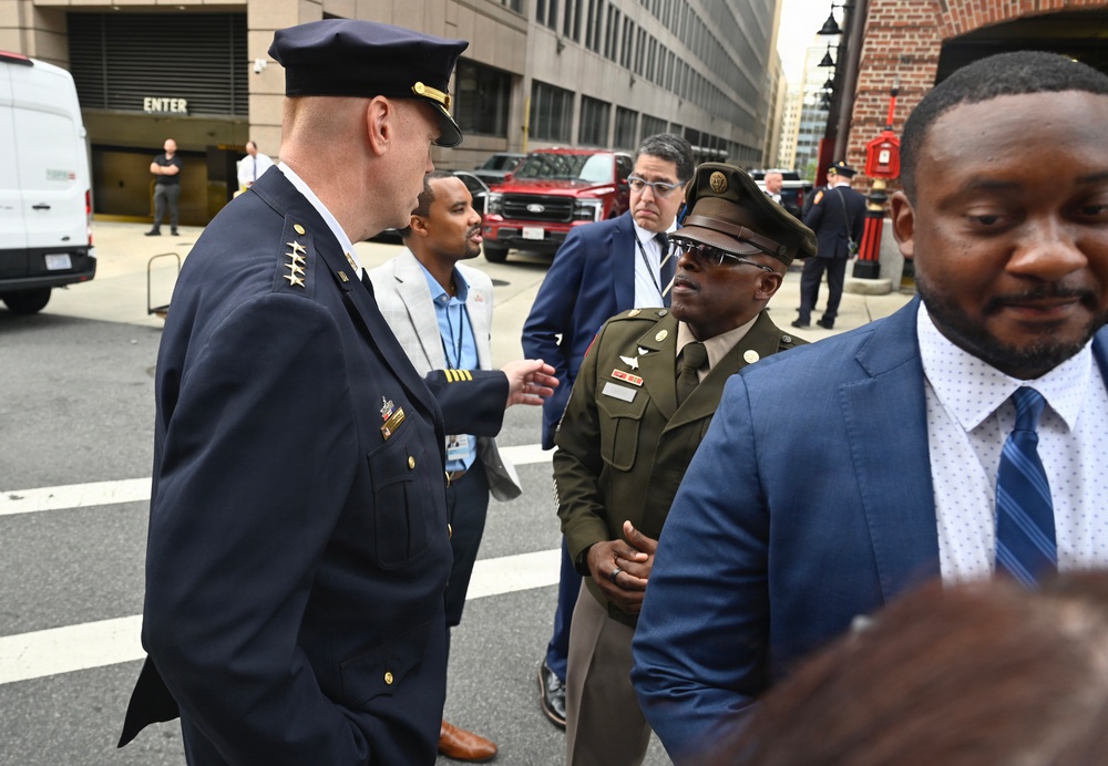 A group of individuals are gathered on a city street, including a person in a dark navy law enforcement dress uniform with gold insignia and a person in an olive/khaki military-style uniform with a garrison cap. Several civilians in business attire are also present in what appears to be an urban setting near a parking garage entrance. The scene suggests an official or formal outdoor gathering or encounter.