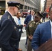 A group of individuals are gathered on a city street, including a person in a dark navy law enforcement dress uniform with gold insignia and a person in an olive/khaki military-style uniform with a garrison cap. Several civilians in business attire are also present in what appears to be an urban setting near a parking garage entrance. The scene suggests an official or formal outdoor gathering or encounter.
