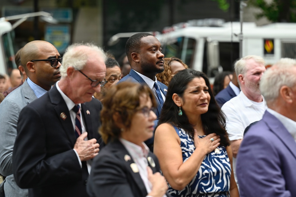 A group of people are gathered outdoors at what appears to be a formal public ceremony or commemoration event. Several individuals in the foreground have their right hands placed over their hearts, suggesting a pledge of allegiance or national anthem moment. The crowd is dressed in business and semi-formal attire, indicating an official or civic occasion.