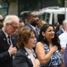 A group of people are gathered outdoors at what appears to be a formal public ceremony or commemoration event. Several individuals in the foreground have their right hands placed over their hearts, suggesting a pledge of allegiance or national anthem moment. The crowd is dressed in business and semi-formal attire, indicating an official or civic occasion.