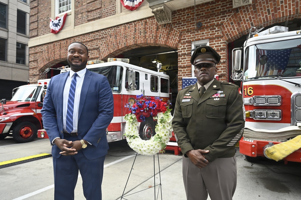Two men stand in front of a fire station beside a memorial wreath decorated with red, white, and blue flowers. One man is dressed in a blue business suit and the other is wearing an olive/green military dress uniform with a peaked cap and service ribbons. The setting appears to be a formal commemorative ceremony, likely honoring fallen first responders or veterans.