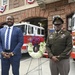Two men stand in front of a fire station beside a memorial wreath decorated with red, white, and blue flowers. One man is dressed in a blue business suit and the other is wearing an olive/green military dress uniform with a peaked cap and service ribbons. The setting appears to be a formal commemorative ceremony, likely honoring fallen first responders or veterans.