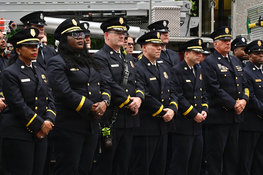 A group of fire department officers in formal dress uniforms are standing at attention in a row, suggesting a ceremonial or memorial event. They are wearing dark navy blue uniforms with gold buttons and yellow sleeve stripes, along with black peaked caps bearing gold badges. A fire truck is visible in the background.