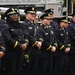 A group of fire department officers in formal dress uniforms are standing at attention in a row, suggesting a ceremonial or memorial event. They are wearing dark navy blue uniforms with gold buttons and yellow sleeve stripes, along with black peaked caps bearing gold badges. A fire truck is visible in the background.