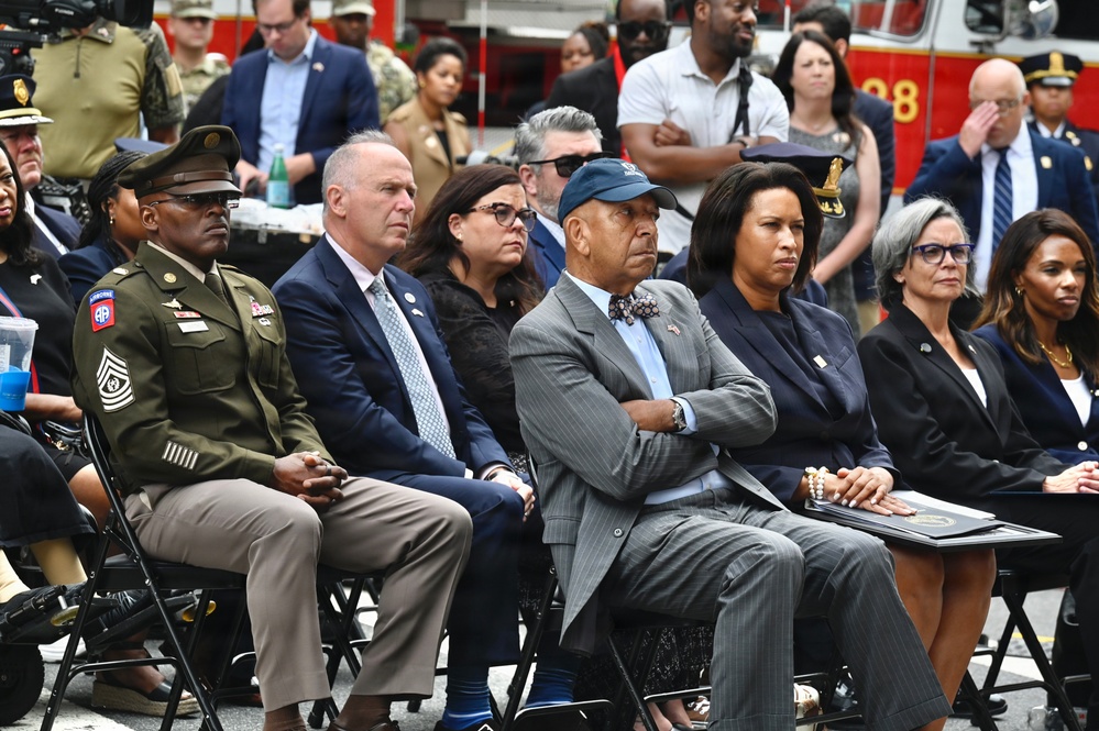 A formal outdoor ceremony or event featuring a diverse group of dignitaries, officials, and military personnel seated in folding chairs. The attendees appear to be observing a presentation or ceremony, with a fire truck visible in the background. The gathering includes military personnel in uniform, civilians in business attire, and what appear to be government officials.