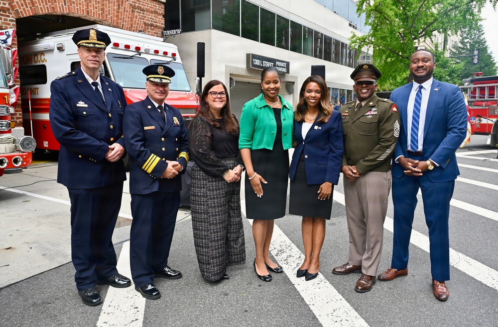 A group of seven individuals pose for a formal photograph on a street in Washington D.C., in front of a DC Fire Department vehicle. The group includes uniformed fire/police officials, military personnel, and civilians in professional attire. The setting appears to be an official or ceremonial event near 1300 L Street.