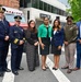 A group of seven individuals pose for a formal photograph on a street in Washington D.C., in front of a DC Fire Department vehicle. The group includes uniformed fire/police officials, military personnel, and civilians in professional attire. The setting appears to be an official or ceremonial event near 1300 L Street.