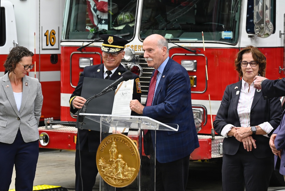An official ceremony is taking place in front of a red fire truck numbered '16', with a podium bearing the District of Columbia seal. A man in a navy suit is presenting or reading from an official document alongside a fire department official in dress uniform. Four individuals are visible, suggesting a formal recognition or proclamation event.