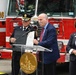 An official ceremony is taking place in front of a red fire truck numbered '16', with a podium bearing the District of Columbia seal. A man in a navy suit is presenting or reading from an official document alongside a fire department official in dress uniform. Four individuals are visible, suggesting a formal recognition or proclamation event.