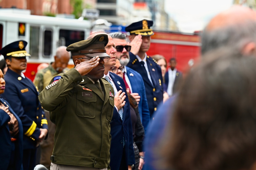 A formal outdoor ceremony featuring military and civilian officials in various uniforms rendering honors, likely during a memorial or commemorative event. A U.S. Army soldier identified by nameplate as 'Smith,' wearing an Airborne tab and olive green uniform, is prominently saluting in the foreground. Behind him, a mix of fire department officials, police officers, military personnel, and civilians in suits are also rendering honors, with a red fire truck visible in the background.