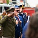 A formal outdoor ceremony featuring military and civilian officials in various uniforms rendering honors, likely during a memorial or commemorative event. A U.S. Army soldier identified by nameplate as 'Smith,' wearing an Airborne tab and olive green uniform, is prominently saluting in the foreground. Behind him, a mix of fire department officials, police officers, military personnel, and civilians in suits are also rendering honors, with a red fire truck visible in the background.