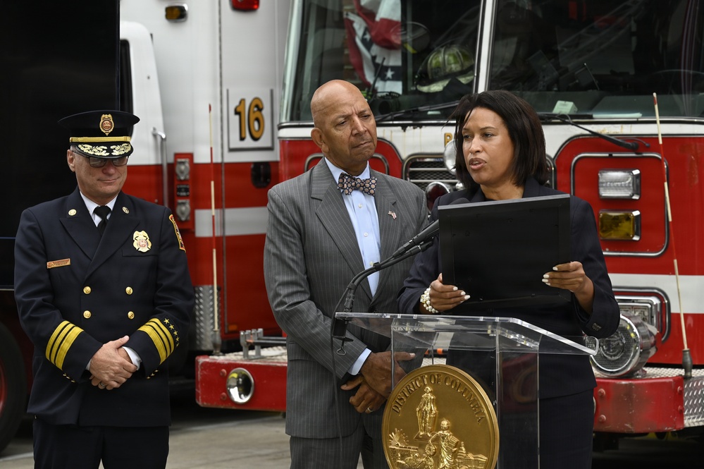 A formal press conference or official ceremony is taking place in front of a red fire truck numbered '16'. A woman speaks at a podium bearing the seal of the District of Columbia, flanked by two men — one in a fire department dress uniform and one in a business suit. The setting appears to be a fire station bay or similar official venue.