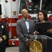 A formal press conference or official ceremony is taking place in front of a red fire truck numbered '16'. A woman speaks at a podium bearing the seal of the District of Columbia, flanked by two men — one in a fire department dress uniform and one in a business suit. The setting appears to be a fire station bay or similar official venue.