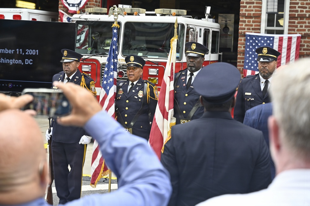 A formal color guard ceremony is taking place in front of a fire station, commemorating the September 11, 2001 attacks in Washington, DC. Four uniformed fire department officers stand at attention, with one holding the American flag and another holding what appears to be the DC flag. Audience members are visible in the foreground, with one person photographing the event.