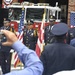 A formal color guard ceremony is taking place in front of a fire station, commemorating the September 11, 2001 attacks in Washington, DC. Four uniformed fire department officers stand at attention, with one holding the American flag and another holding what appears to be the DC flag. Audience members are visible in the foreground, with one person photographing the event.