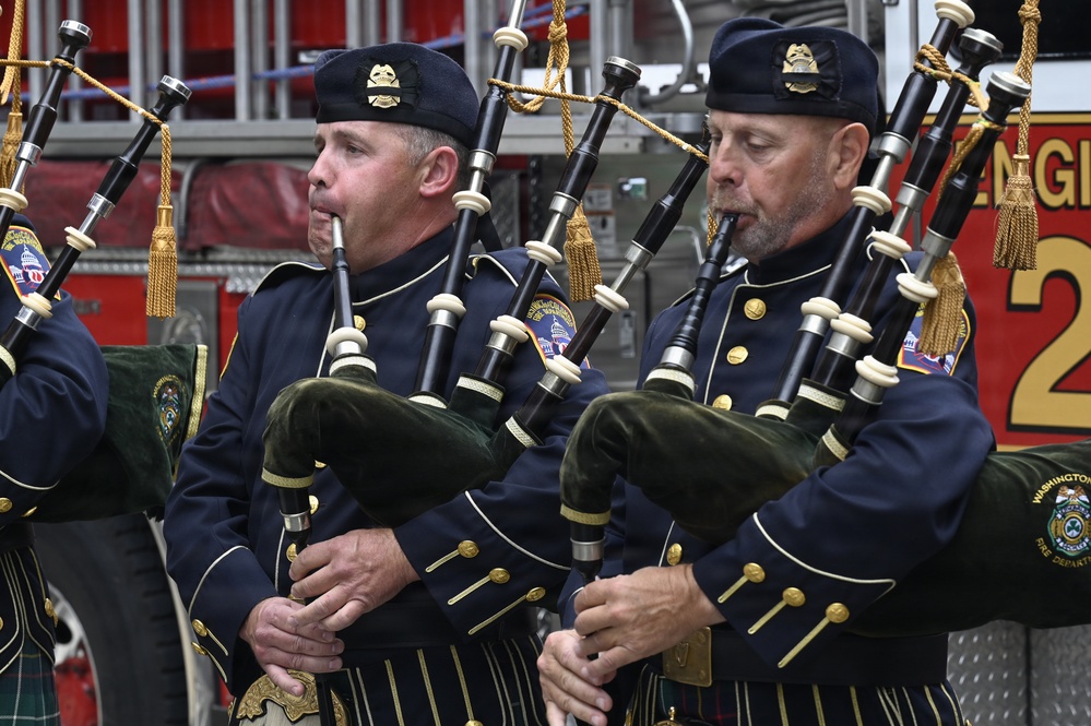 Two uniformed bagpipers are playing bagpipes in front of a red fire engine. They are wearing dark navy dress uniforms with gold trim and flat caps bearing badges, consistent with a fire department pipe and drum band. Patches and text visible on their uniforms reference the Washington Fire Department.