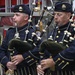 Two uniformed bagpipers are playing bagpipes in front of a red fire engine. They are wearing dark navy dress uniforms with gold trim and flat caps bearing badges, consistent with a fire department pipe and drum band. Patches and text visible on their uniforms reference the Washington Fire Department.