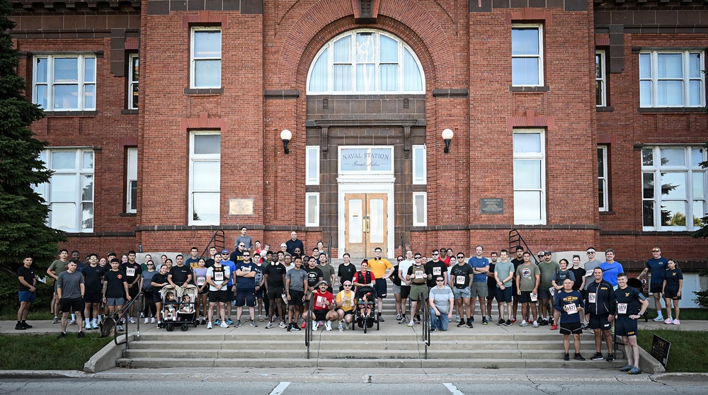 A large group of approximately 80-100 people poses for a group photo on the steps of Naval Station Great Lakes, a historic red brick building. The gathering appears to be associated with a fun run or 5K event, as many participants are wearing numbered race bibs. The group includes military personnel and civilians of diverse ages, with some participants using wheelchairs and strollers.