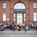 A large group of approximately 80-100 people poses for a group photo on the steps of Naval Station Great Lakes, a historic red brick building. The gathering appears to be associated with a fun run or 5K event, as many participants are wearing numbered race bibs. The group includes military personnel and civilians of diverse ages, with some participants using wheelchairs and strollers.