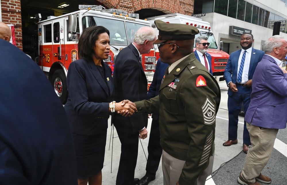 A formal outdoor event taking place in front of a fire station, featuring Engine Company 16. A woman in a dark business suit is shaking hands with a U.S. Army soldier in dress uniform, while several other officials and attendees stand nearby. The setting appears to be a public ceremony or official visit.