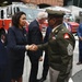 A formal outdoor event taking place in front of a fire station, featuring Engine Company 16. A woman in a dark business suit is shaking hands with a U.S. Army soldier in dress uniform, while several other officials and attendees stand nearby. The setting appears to be a public ceremony or official visit.
