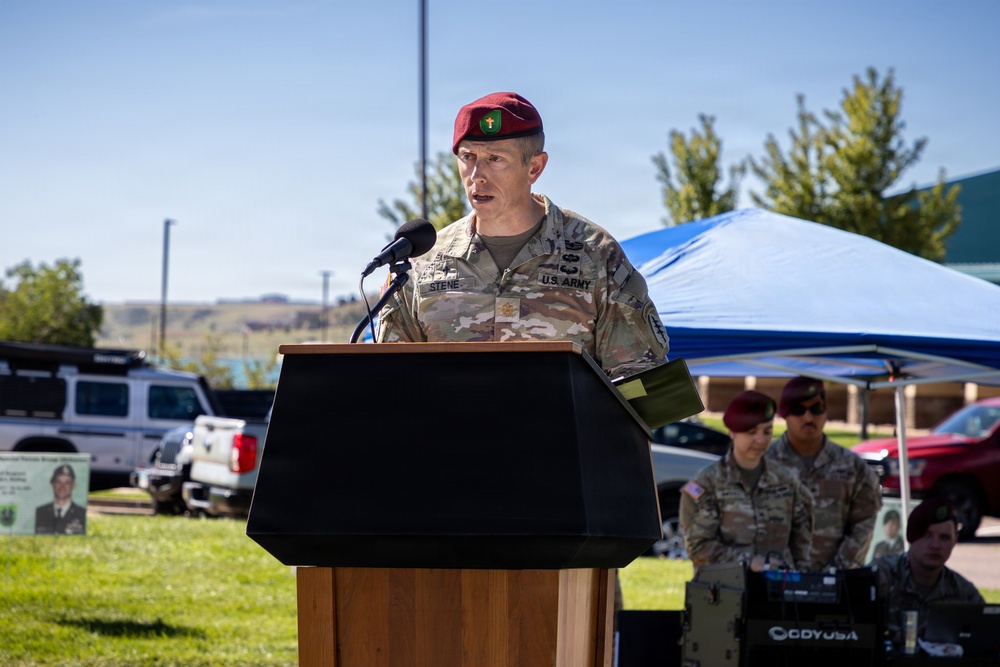 A U.S. Army soldier wearing a maroon beret and multicam uniform is speaking at a wooden podium during an outdoor military ceremony. The name tape on the uniform reads 'STENE' and the soldier wears a U.S. Army branch tape. Several other soldiers in similar uniforms, some also wearing maroon berets, are visible in the background under a blue canopy tent.