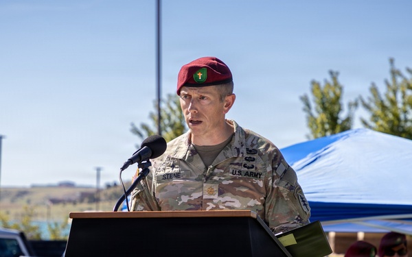 A U.S. Army soldier wearing a maroon beret and multicam uniform is speaking at a wooden podium during an outdoor military ceremony. The name tape on the uniform reads 'STENE' and the soldier wears a U.S. Army branch tape. Several other soldiers in similar uniforms, some also wearing maroon berets, are visible in the background under a blue canopy tent.