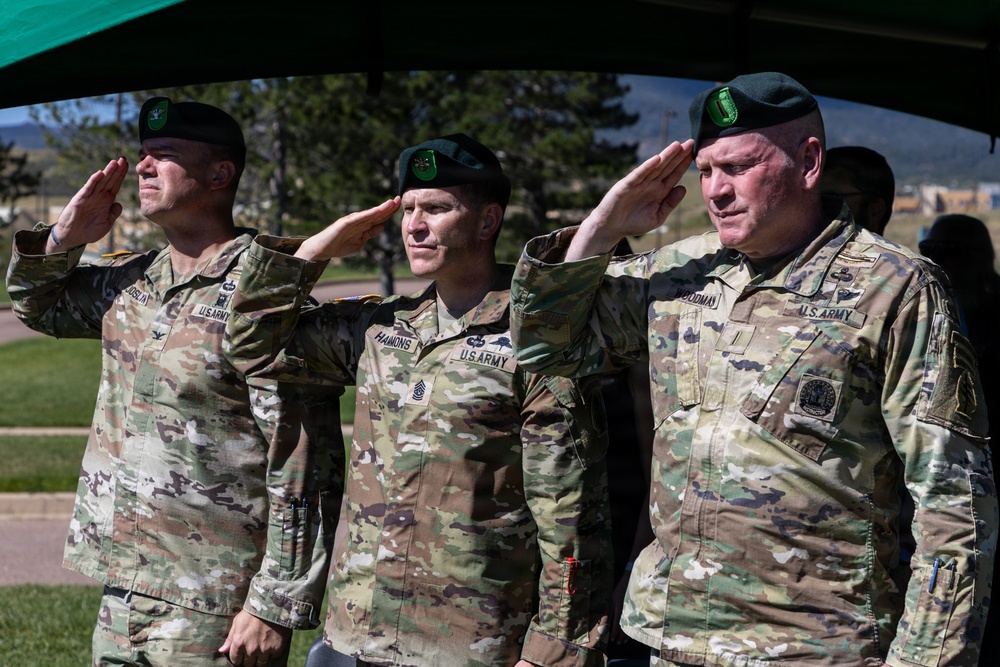 Three U.S. Army soldiers in OCP (Operational Camouflage Pattern) uniforms and green berets are rendering a salute during what appears to be a formal military ceremony. Name tapes visible on the uniforms read 'HAMMONS' (center) and 'GOODMAN' (right), with a partially visible name on the left. The setting appears to be an outdoor military installation with trees and buildings visible in the background.