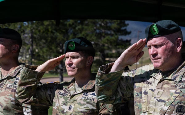 Three U.S. Army soldiers in OCP (Operational Camouflage Pattern) uniforms and green berets are rendering a salute during what appears to be a formal military ceremony. Name tapes visible on the uniforms read 'HAMMONS' (center) and 'GOODMAN' (right), with a partially visible name on the left. The setting appears to be an outdoor military installation with trees and buildings visible in the background.
