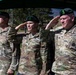 Three U.S. Army soldiers in OCP (Operational Camouflage Pattern) uniforms and green berets are rendering a salute during what appears to be a formal military ceremony. Name tapes visible on the uniforms read 'HAMMONS' (center) and 'GOODMAN' (right), with a partially visible name on the left. The setting appears to be an outdoor military installation with trees and buildings visible in the background.
