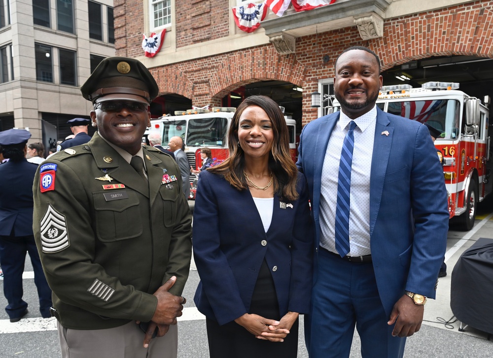 Three individuals pose for a photo in front of a fire station decorated with patriotic red, white, and blue bunting. The group includes a U.S. Army soldier in dress uniform, a woman in a navy blazer, and a man in a blue suit with a striped tie. The setting suggests a patriotic or military commemorative event.