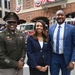 Three individuals pose for a photo in front of a fire station decorated with patriotic red, white, and blue bunting. The group includes a U.S. Army soldier in dress uniform, a woman in a navy blazer, and a man in a blue suit with a striped tie. The setting suggests a patriotic or military commemorative event.