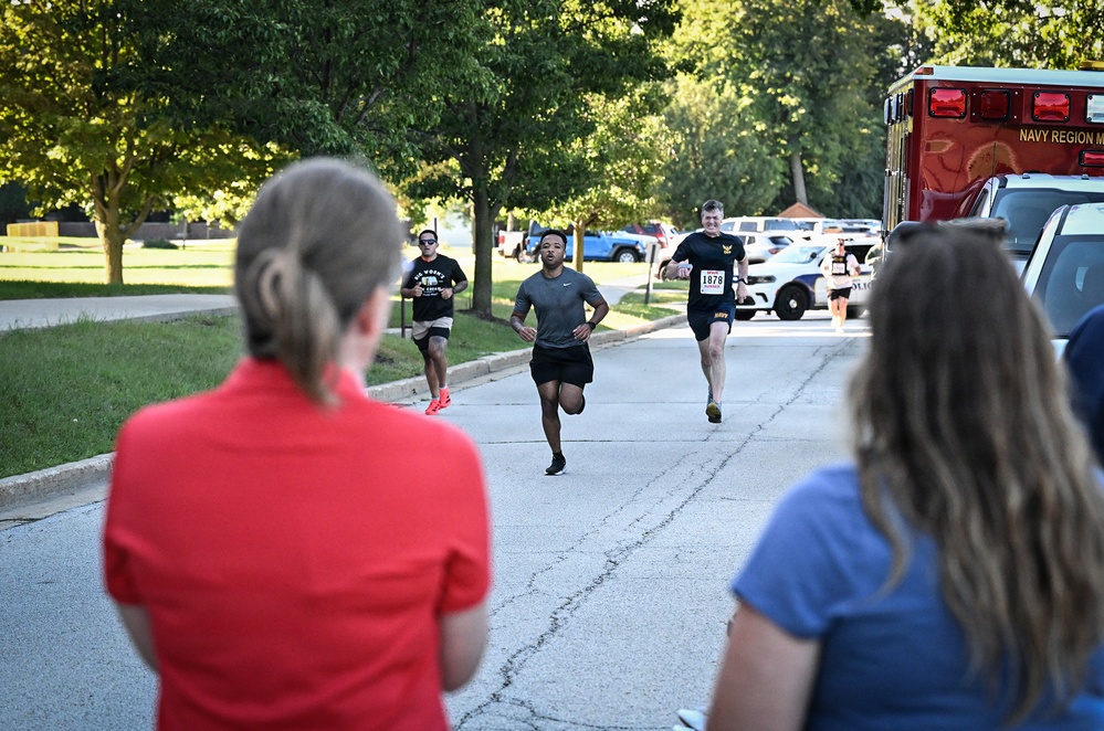 A road race or fun run event is taking place on a paved road, likely on a military installation given the visible 'Navy Region M' ambulance and Navy-branded race bib. Several runners are approaching the finish area while two spectators in the foreground watch with their backs to the camera. The setting includes green trees, parked vehicles, and what appears to be a police vehicle in the background.