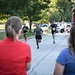 A road race or fun run event is taking place on a paved road, likely on a military installation given the visible 'Navy Region M' ambulance and Navy-branded race bib. Several runners are approaching the finish area while two spectators in the foreground watch with their backs to the camera. The setting includes green trees, parked vehicles, and what appears to be a police vehicle in the background.