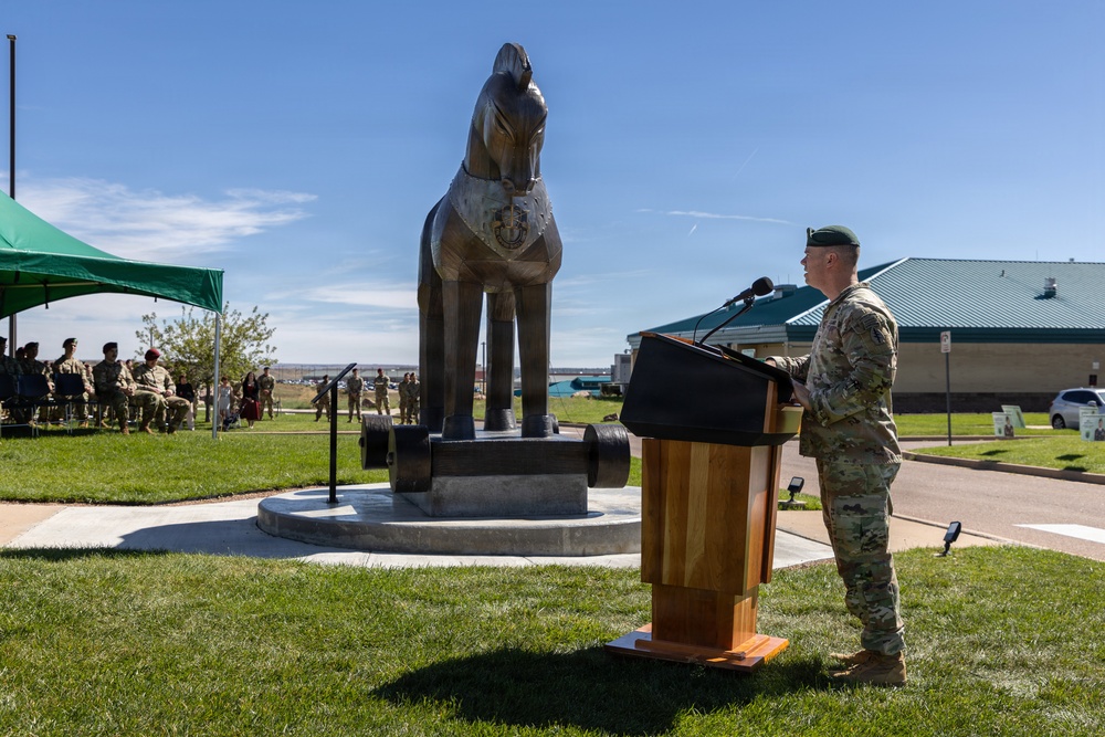 A military officer wearing a green beret and camouflage uniform speaks at a wooden podium during an outdoor ceremony. Behind him stands a large bronze horse statue mounted on a circular stone pedestal, likely a unit or installation monument. An audience of military personnel and civilians is seated under a green canopy tent to the left.