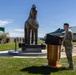 A military officer wearing a green beret and camouflage uniform speaks at a wooden podium during an outdoor ceremony. Behind him stands a large bronze horse statue mounted on a circular stone pedestal, likely a unit or installation monument. An audience of military personnel and civilians is seated under a green canopy tent to the left.